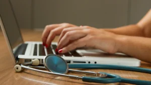 Medical professional typing on a laptop beside a stethoscope on a wooden desk