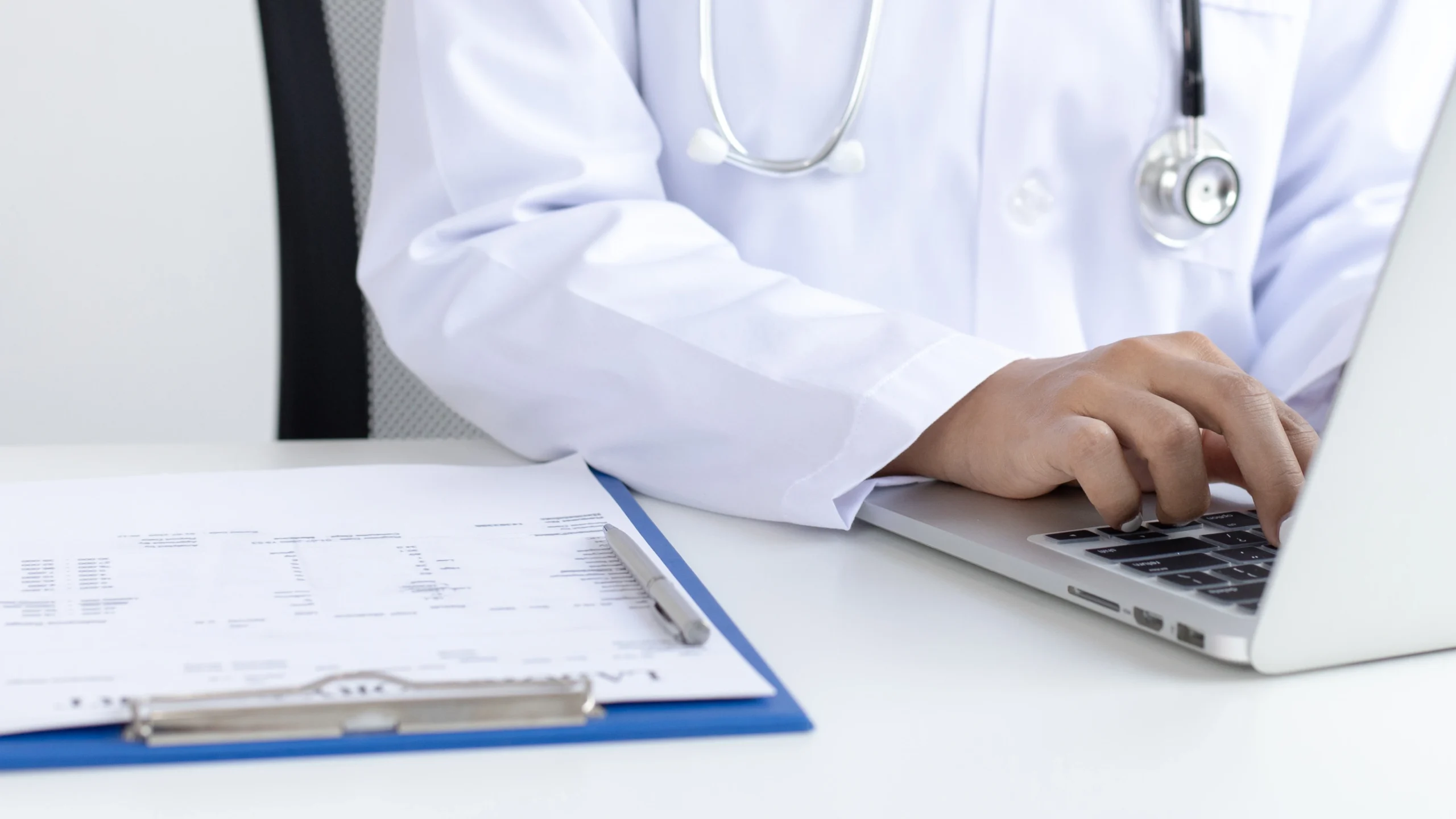 Physician in a white coat entering codes on a laptop next to a printed medical record on a clipboard