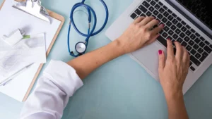 Clinician in a white coat working at a laptop beside a clipboard and stethoscope