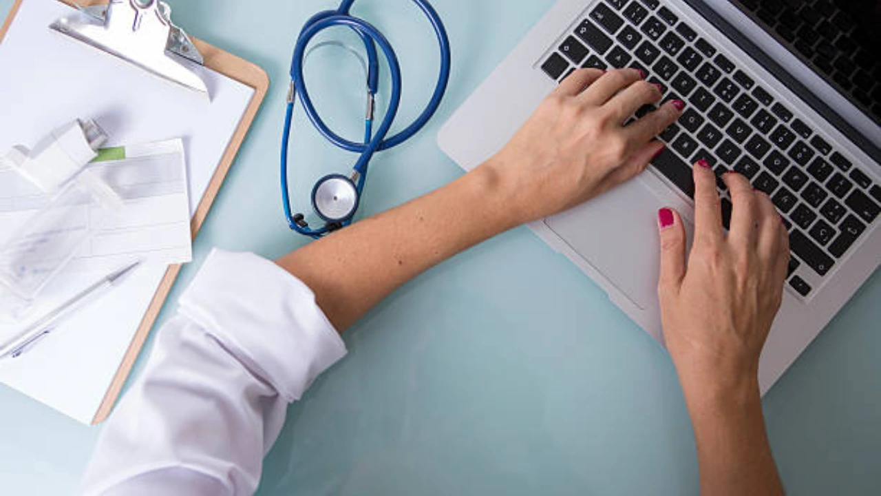 Clinician in a white coat working at a laptop beside a clipboard and stethoscope