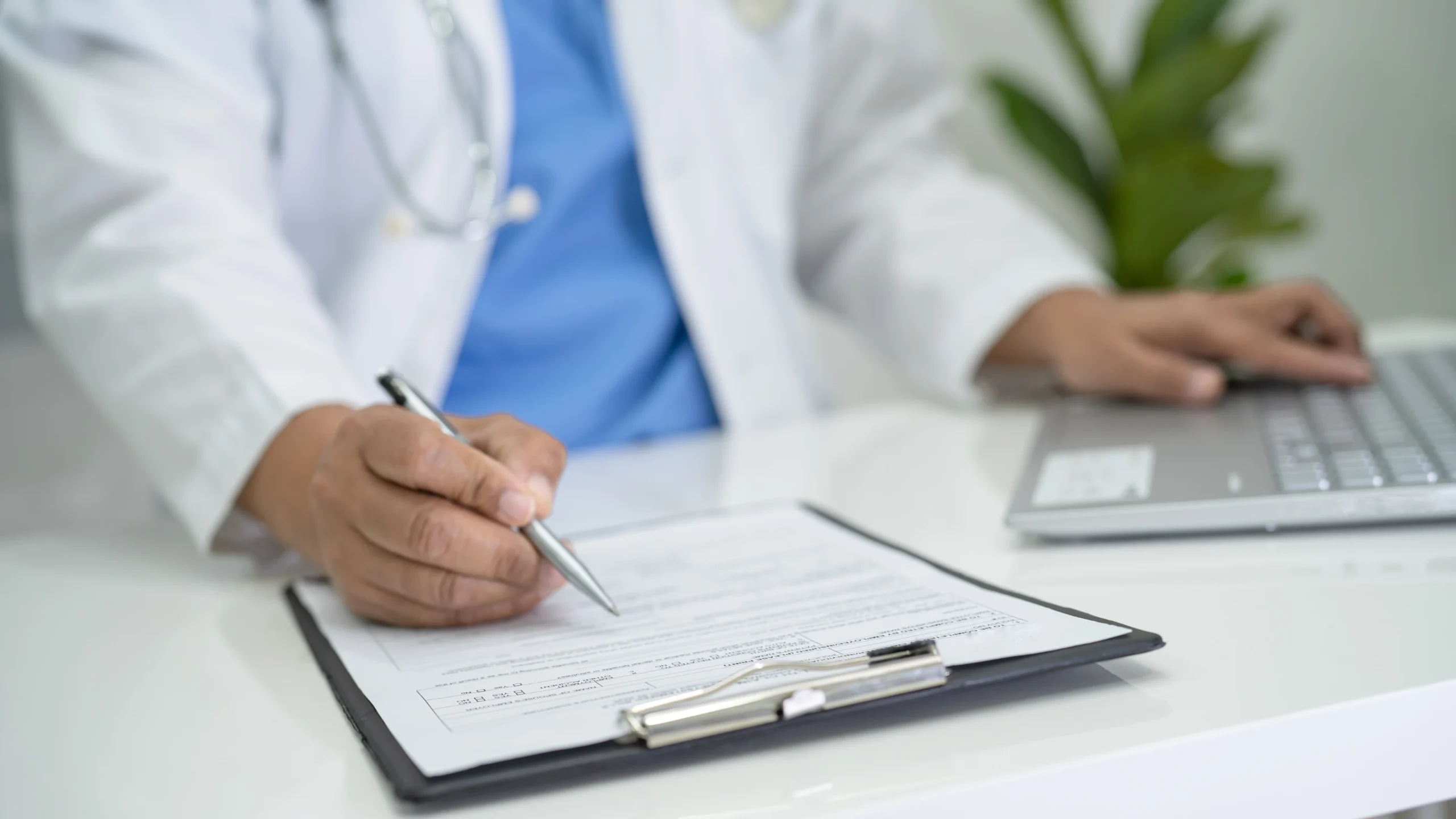 Physician in a white coat completing a claim form on a clipboard beside a laptop