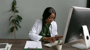 Physician in a white coat entering billing information at a desktop computer beside a clipboard