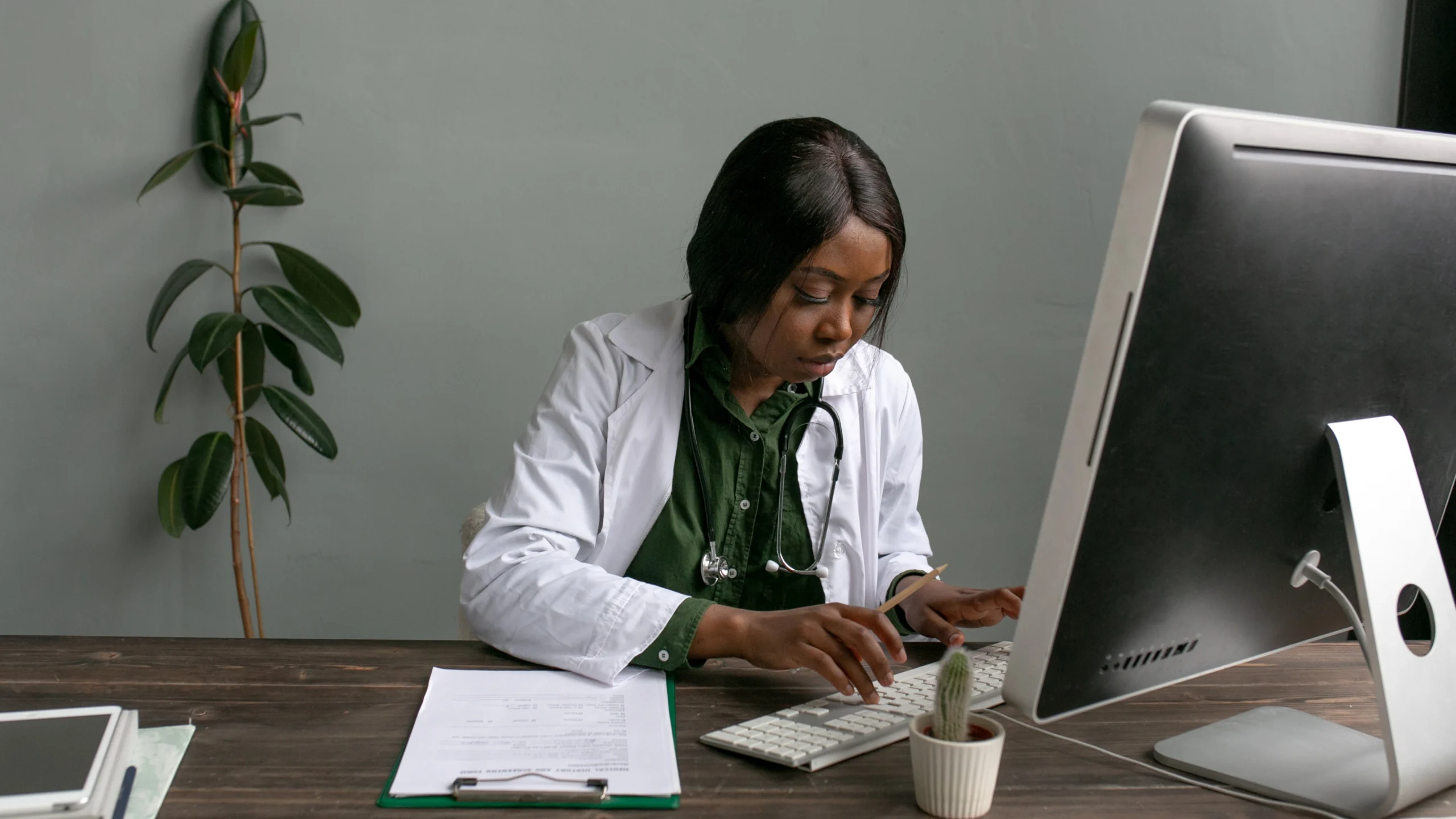 Physician in a white coat entering billing information at a desktop computer beside a clipboard
